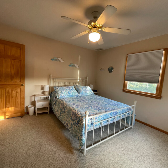 Bedroom with a white metal bed frame, blue bedding, ceiling fan, nightstands, and a window with blinds.