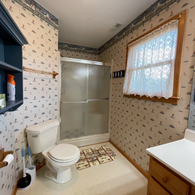 Bathroom with patterned wallpaper, toilet, glass shower, lace-curtained window, and vanity with sink and mirror.