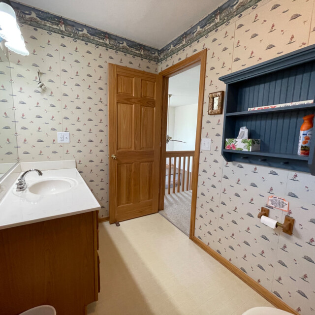 Bathroom with nautical wallpaper, wooden vanity, mirror, wooden door, and blue wall shelf above the toilet paper holder.