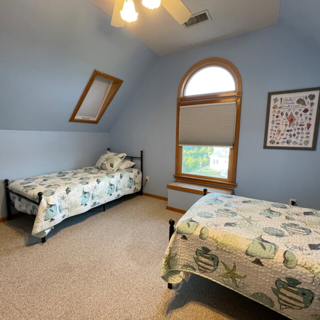 Bedroom with two twin beds, blue walls, a window with a shade, framed art, and patterned bedding.