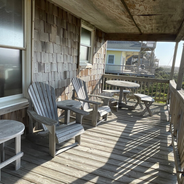Sunny wooden porch with Adirondack chairs, side tables, and a picnic table, overlooking greenery and nearby houses.