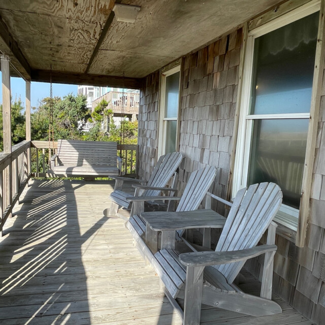 Wooden porch with two Adirondack chairs, a small table, and a porch swing facing trees and nearby houses.