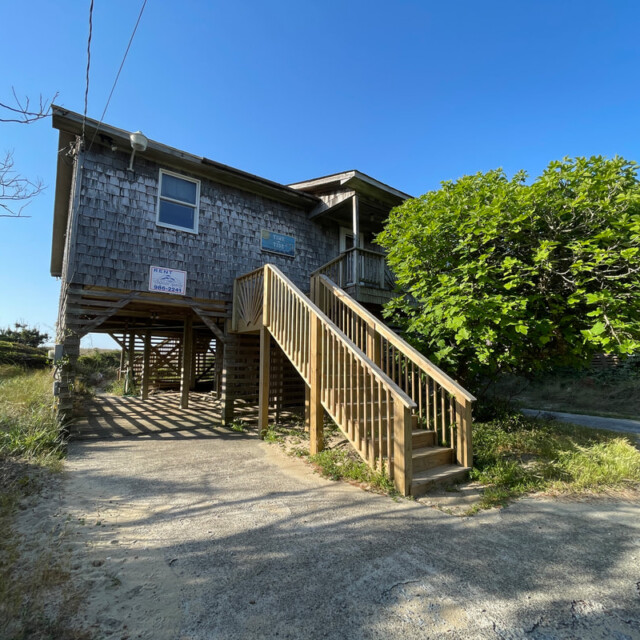 A wooden beach house on stilts with an exterior staircase, surrounded by greenery under a clear blue sky.