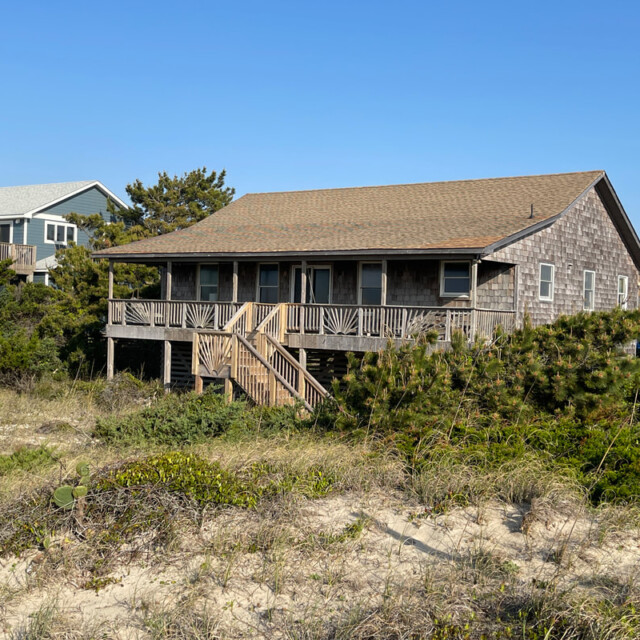 Wooden beach house with a wraparound porch, surrounded by sand dunes and greenery under a clear blue sky.