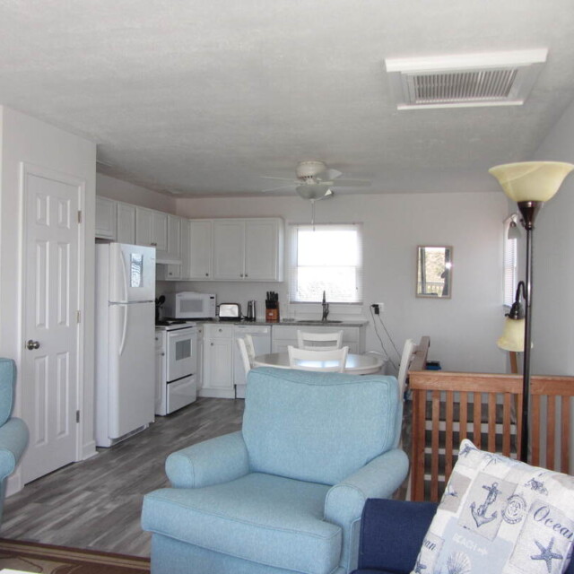 Open living area with blue armchairs, white kitchen, dining table, and a floor lamp near a window with blinds.