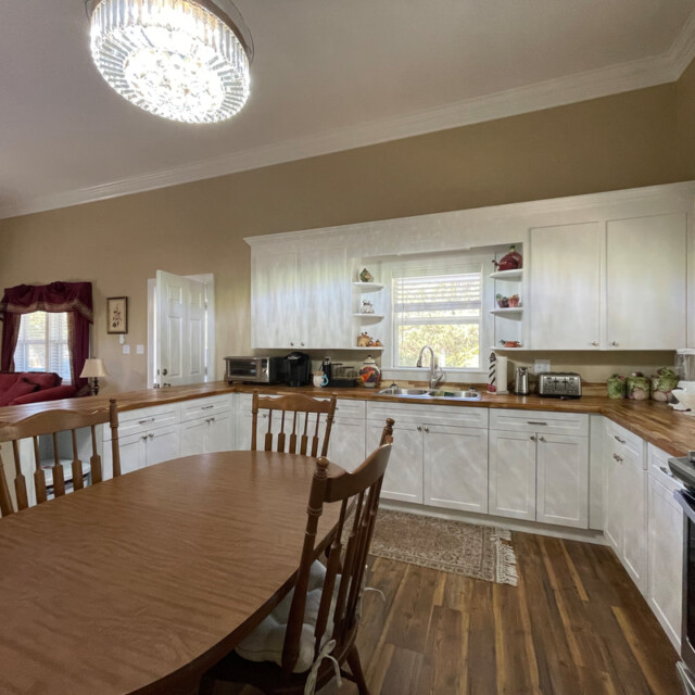 Spacious kitchen with wooden dining table, white cabinets, large window, and stainless steel appliances.