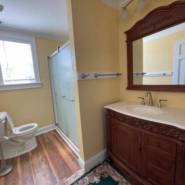 Bathroom with wood vanity, large mirror, toilet, glass shower, yellow walls, and wood-look flooring.