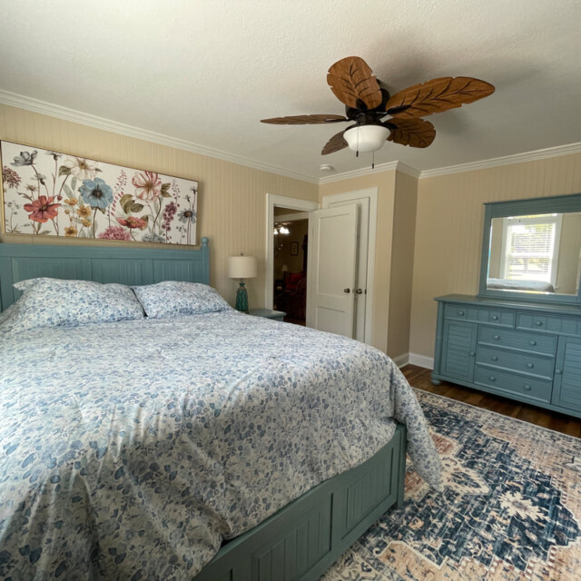 Bedroom with a blue bed and dresser, floral wall art, two lamps, a ceiling fan, and a patterned rug.
