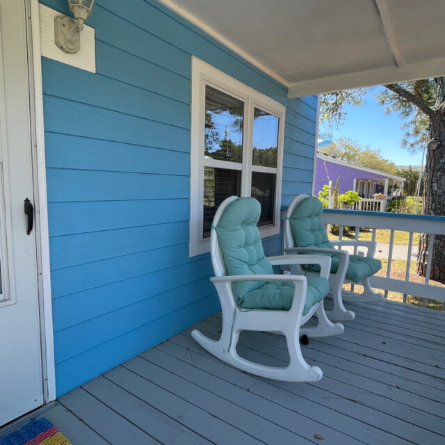 Two padded rocking chairs sit on a blue-painted porch next to a white door and a window, with trees in the background.