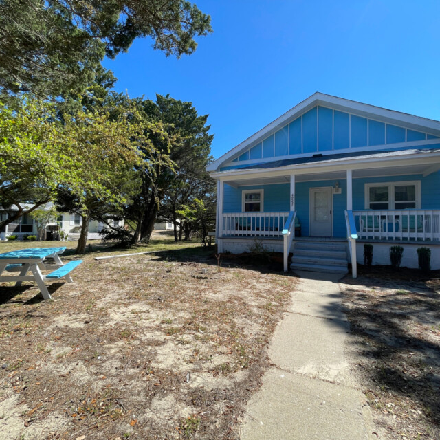A small blue house with a front porch and a picnic table in a yard with sparse grass and trees.
