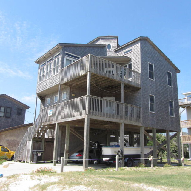 Three-story beach house on stilts with wooden decks, cars, and a boat parked underneath, set on a sunny day.