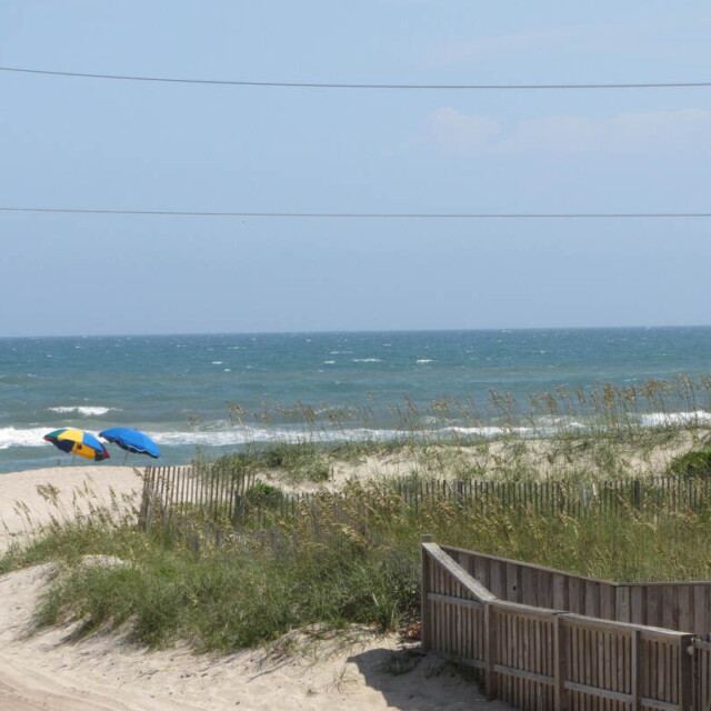 Wooden fence and grassy sand dunes lead to a beach with two umbrellas near the shoreline under a partly cloudy sky.