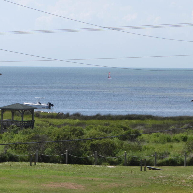 View of grassy dunes and buildings near the shore with a boat and a distant windsurfer on the calm sea under a clear sky.