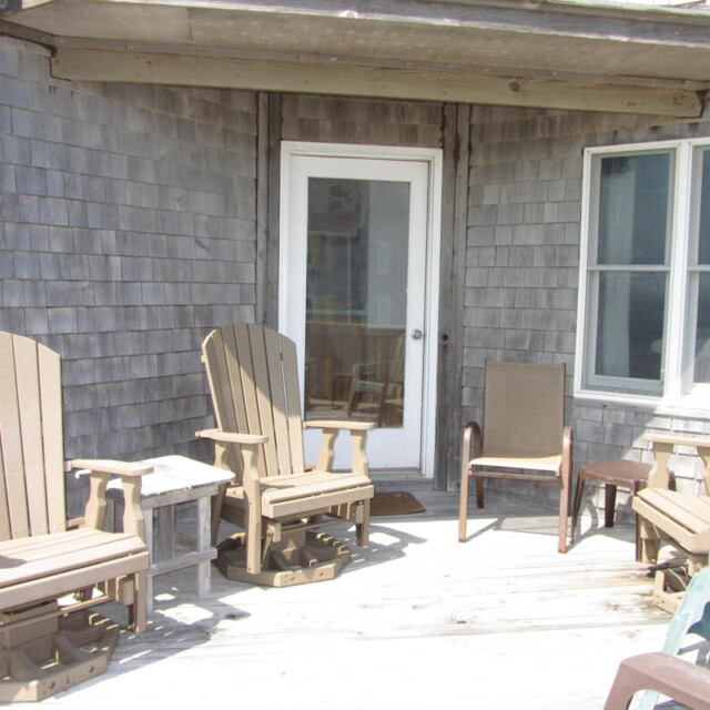 Four wooden chairs and a small table are arranged on a sunlit wooden deck outside a house with shingle siding.