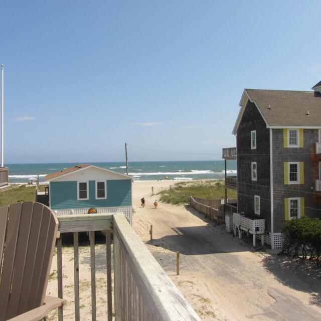 View of a sandy road leading to the beach, flanked by two houses, with the ocean visible in the background.