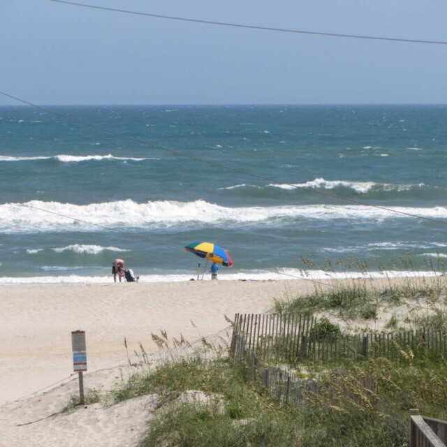 Two people with a colorful umbrella stand on a sandy beach near the ocean, with waves and sea grass visible.