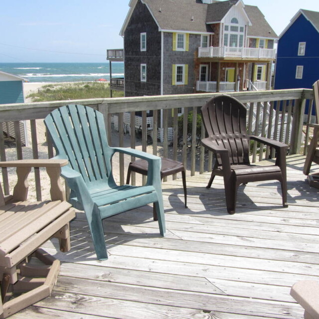 Several Adirondack chairs on a wooden deck overlook beach houses and the ocean under a partly cloudy sky.