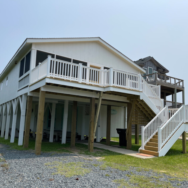 White elevated beach house with a large deck, wooden stairs, and gravel driveway on a sunny day.
