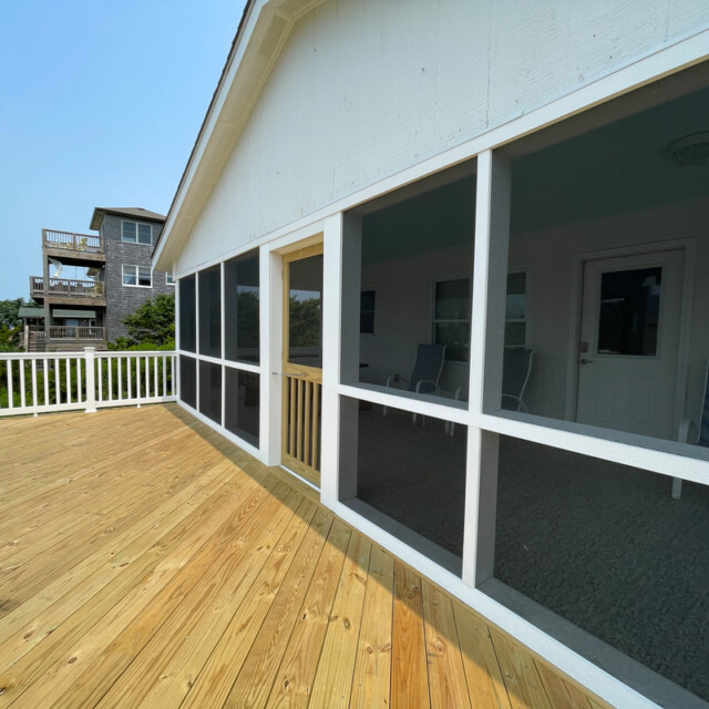 Wooden deck with white railing next to a screened-in porch attached to a white house on a sunny day.
