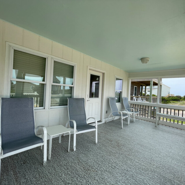 Covered porch with four chairs, a small table, windows, and doors; view of another house and greenery outside.