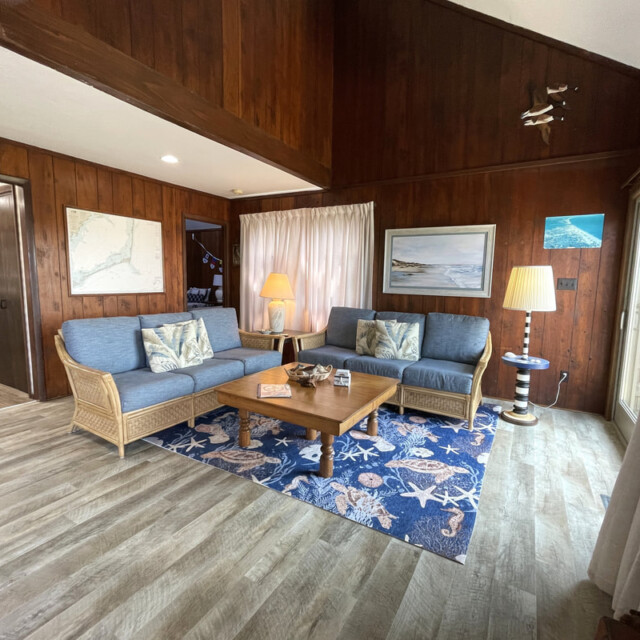 Living room with wood paneling, blue sofas, nautical rug, two lamps, coffee table, and large window with light curtains.
