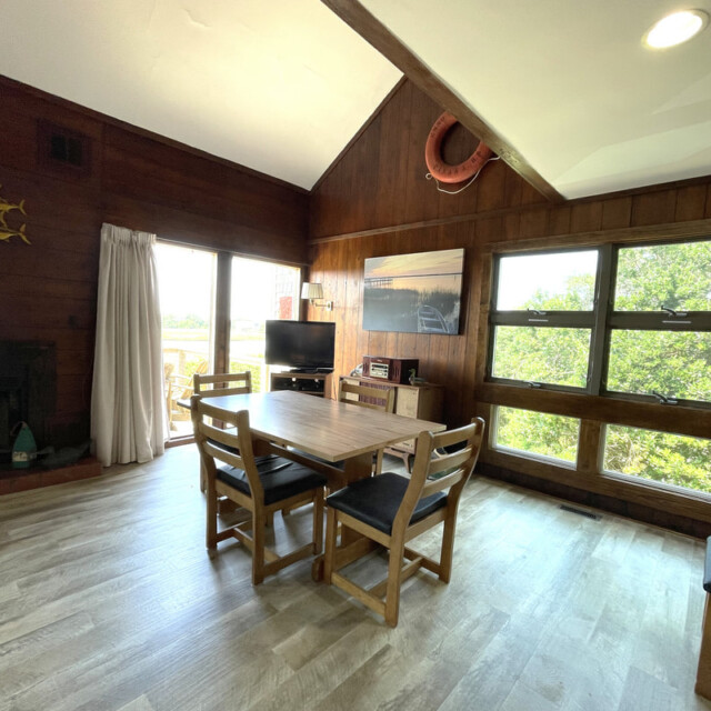 Dining table with four chairs in a wood-paneled room, large windows, and a TV on a stand near a sliding glass door.