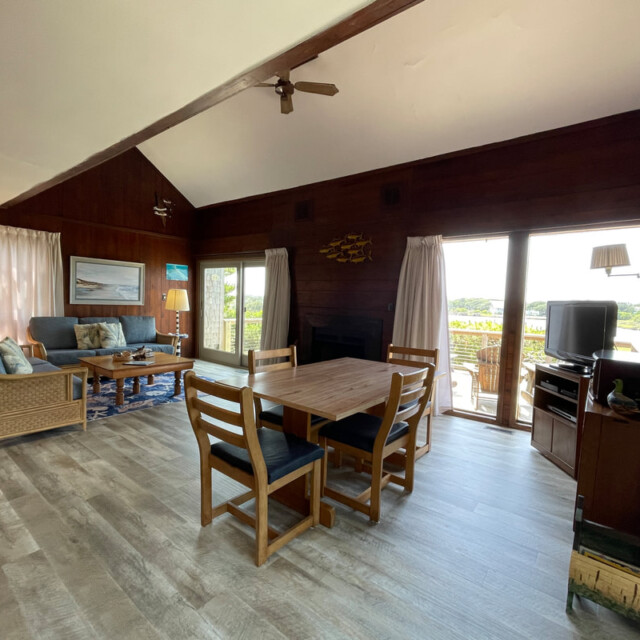 Wood-paneled living room with dining table, sofas, TV, and large windows overlooking a deck and outdoor view.