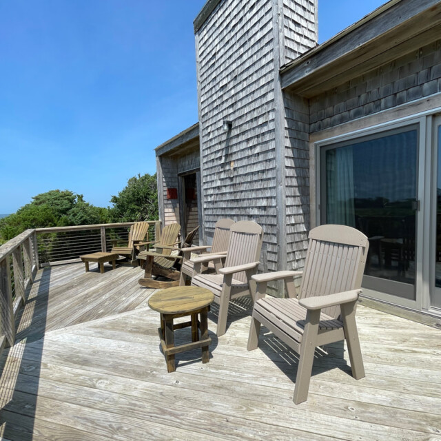 Wooden deck with Adirondack chairs and a small table, overlooking greenery and water under a clear blue sky.