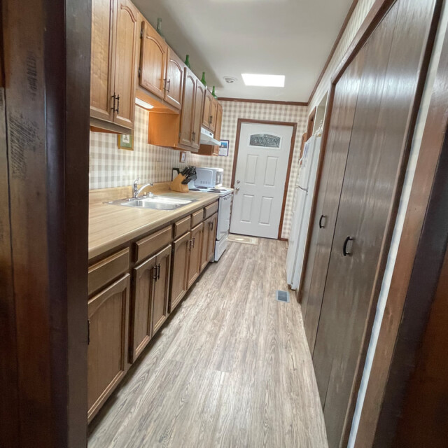 Narrow kitchen with wooden cabinets, laminate floor, and a white door at the far end, seen from the hallway.