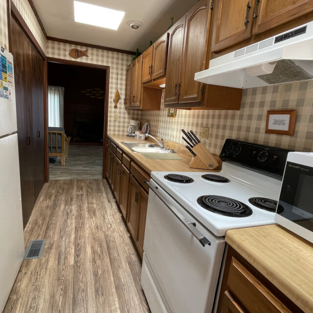 Narrow kitchen with wood cabinets, white appliances, microwave, and wood-patterned flooring under bright sunlight.