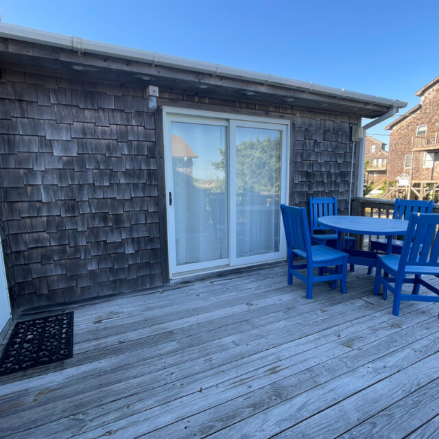 Wooden deck with a round table and four blue chairs beside a sliding glass door and shingled exterior wall.