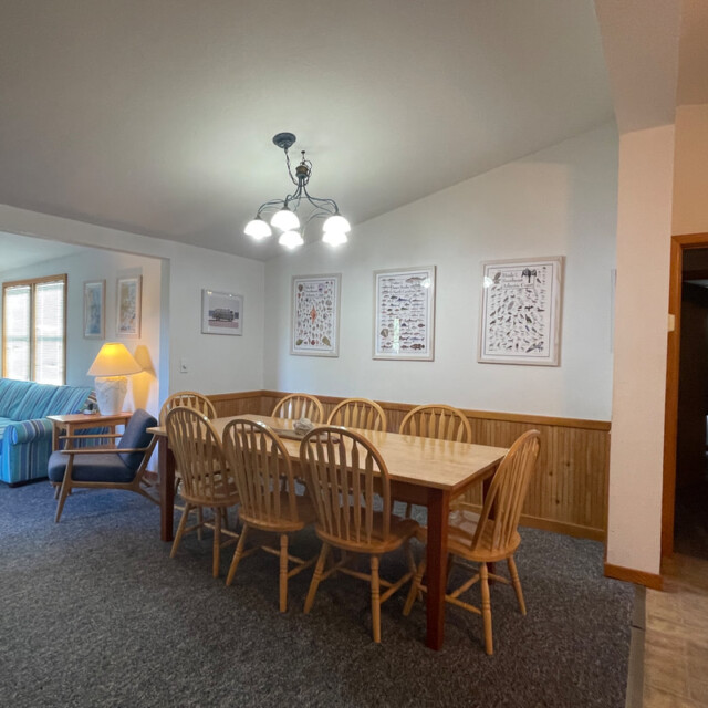 Dining area with a wooden table and eight chairs, adjacent to a living room with a sofa and windows.