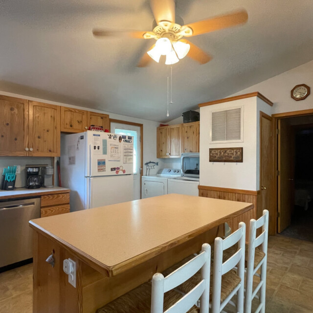 Kitchen with wooden cabinets, white appliances, island with three chairs, and a ceiling fan with light above.