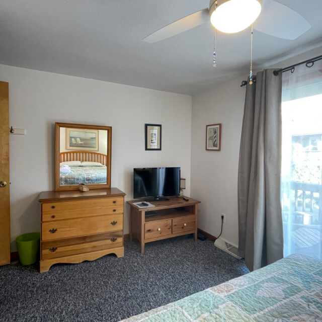 Bedroom with dresser, mirror, TV on a stand, bed partially visible, window with sheer curtains, and ceiling fan.