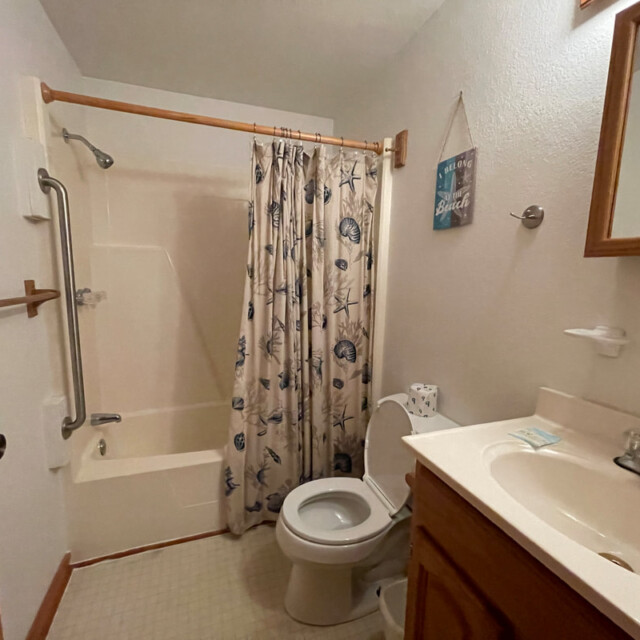 Small bathroom with a shower-tub combo, curtain with sea motifs, toilet, sink, and wooden accents.