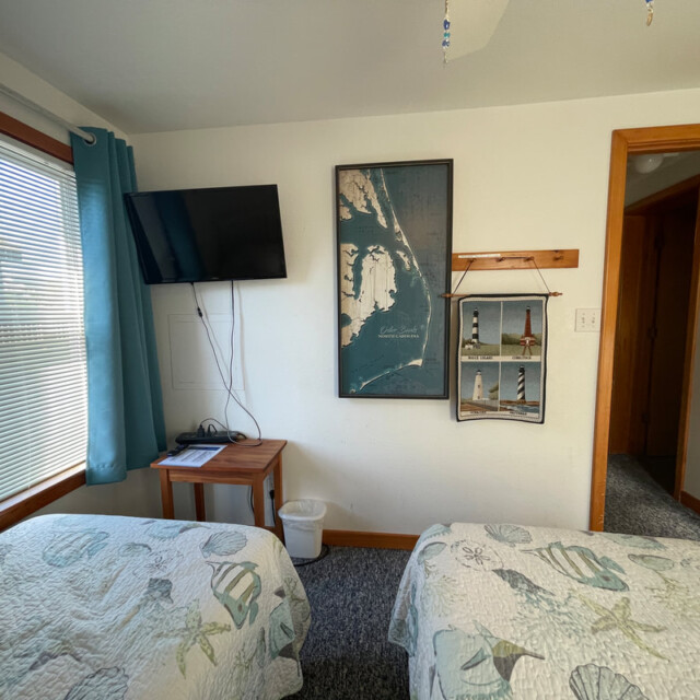 Small bedroom with two twin beds, wall art, a mounted TV, and sunlight streaming through a window with blue curtains.