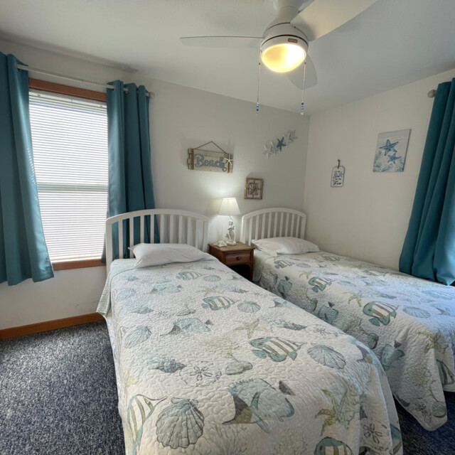 Bedroom with two twin beds, seashell bedding, blue curtains, and beach-themed wall decor. Light on nightstand is on.