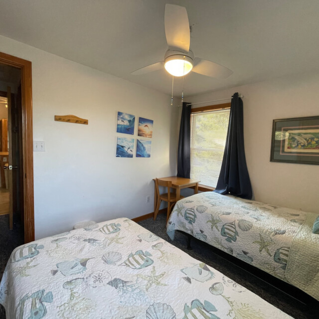 Bedroom with two twin beds, seashell-themed bedding, a small desk by the window, and wall art.