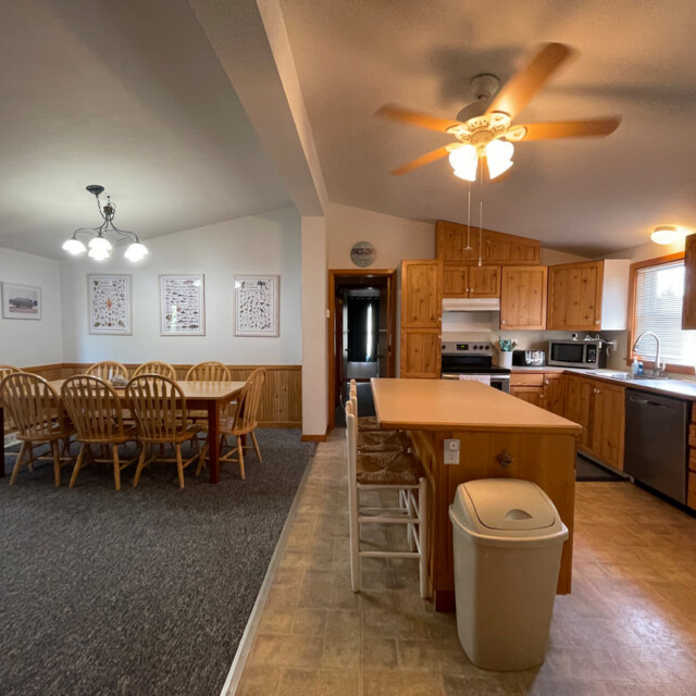 Open kitchen and dining area with wooden cabinets, a dining table for eight, ceiling fans, and bright lighting.