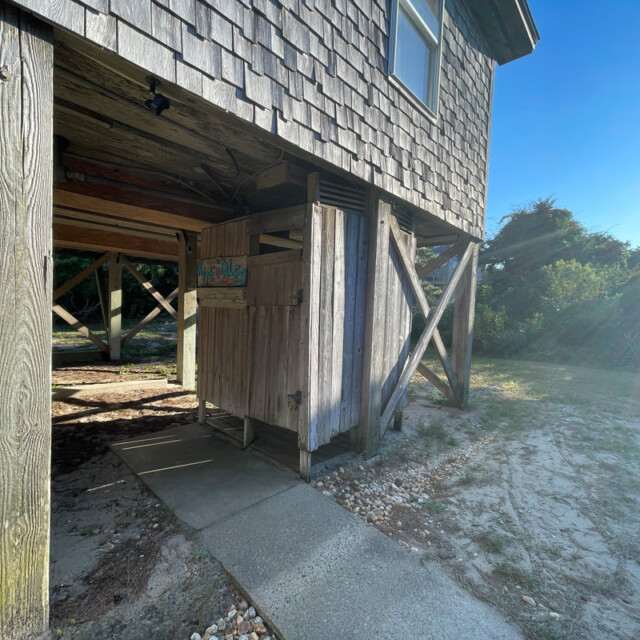 Wooden outdoor shower under a raised house, with sunlight casting shadows and trees in the background.