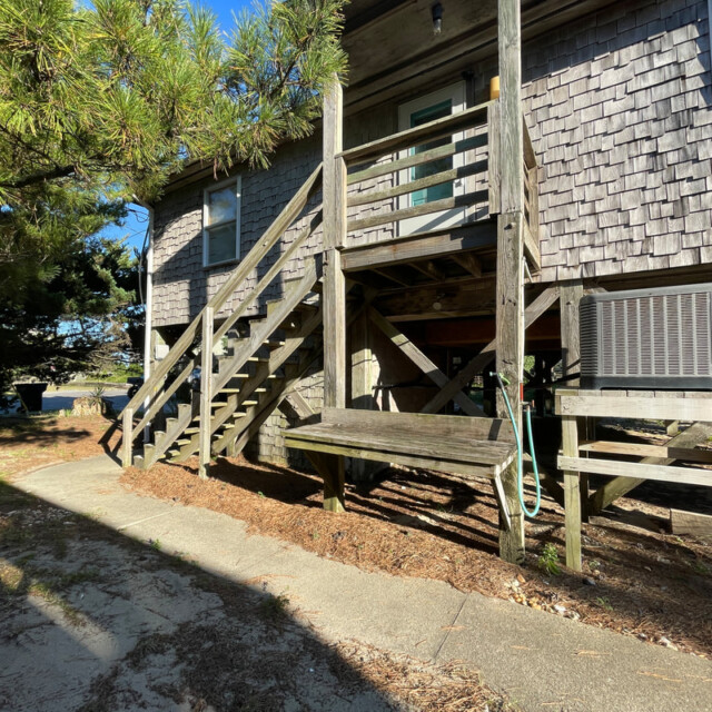 Wooden stairs lead up to a shingled house with an air conditioning unit and hose underneath. Sunlight casts shadows.