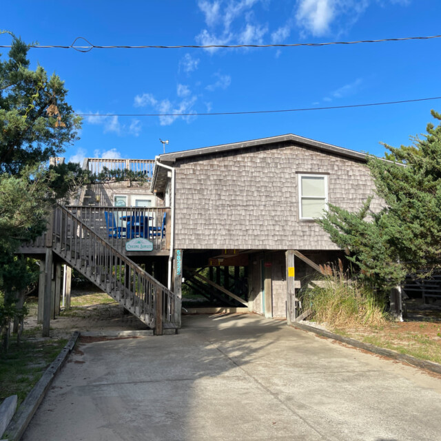 Wooden beach house on stilts with a long driveway, surrounded by trees and clear blue sky.