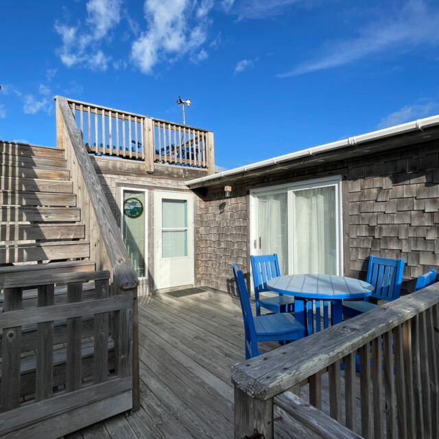 Wooden deck with blue table and chairs, staircase leading to rooftop, and sliding glass door under a clear blue sky.