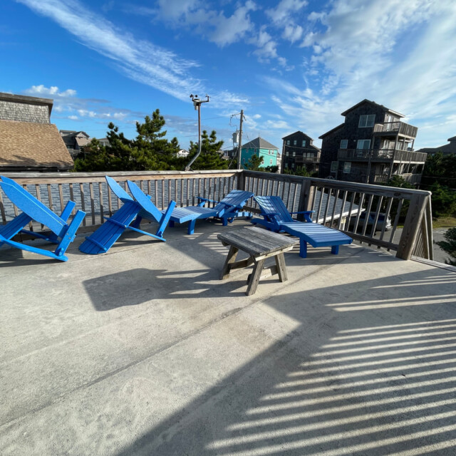 Rooftop deck with several blue wooden lounge chairs and a small bench, surrounded by a wooden railing, under a blue sky.