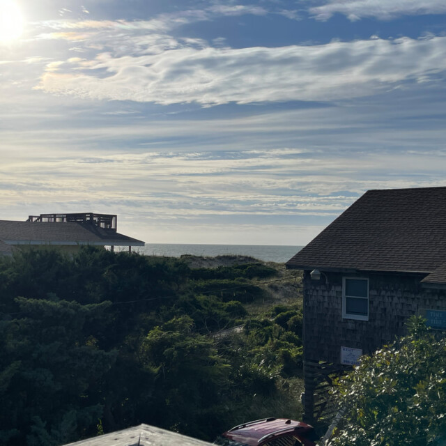 Sunlight shines over beach houses, greenery, and the ocean under a partly cloudy sky in the early morning.