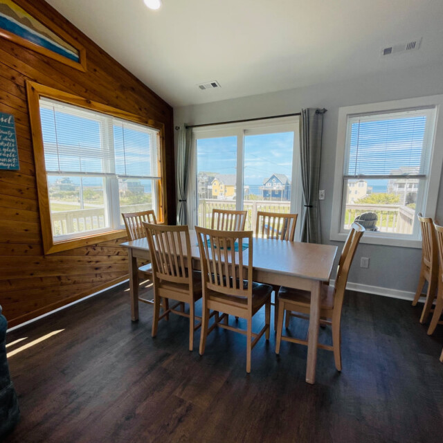 Dining area with a wooden table and chairs, large windows, and views of houses and sky outside.