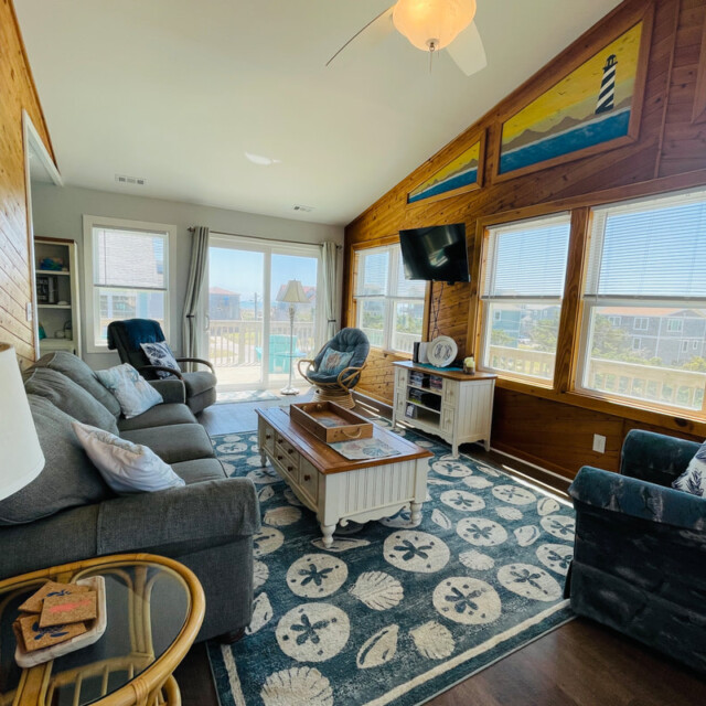Bright living room with wood walls, coastal decor, large windows, gray sofas, and a view of houses outside.