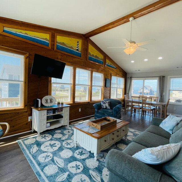 Bright living room with large windows, coastal decor, wood accents, and views of houses and the ocean outside.