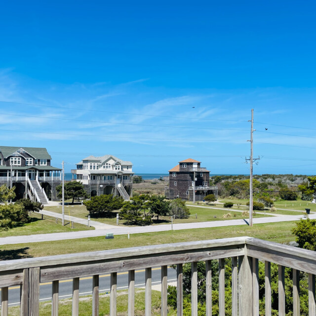 View from a wooden deck overlooking houses, grassy yards, and a distant ocean under a clear blue sky.