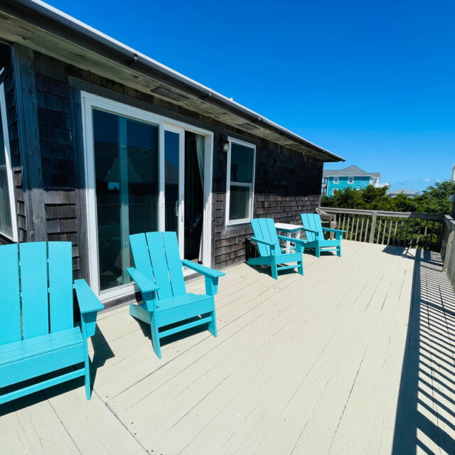 Four turquoise Adirondack chairs on a spacious wooden deck outside a dark-shingled house under a clear blue sky.
